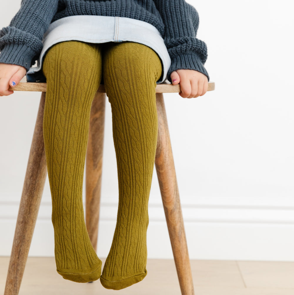 little girl sitting on wooden stool wearing bright olive green cable knit tights under denim skirt