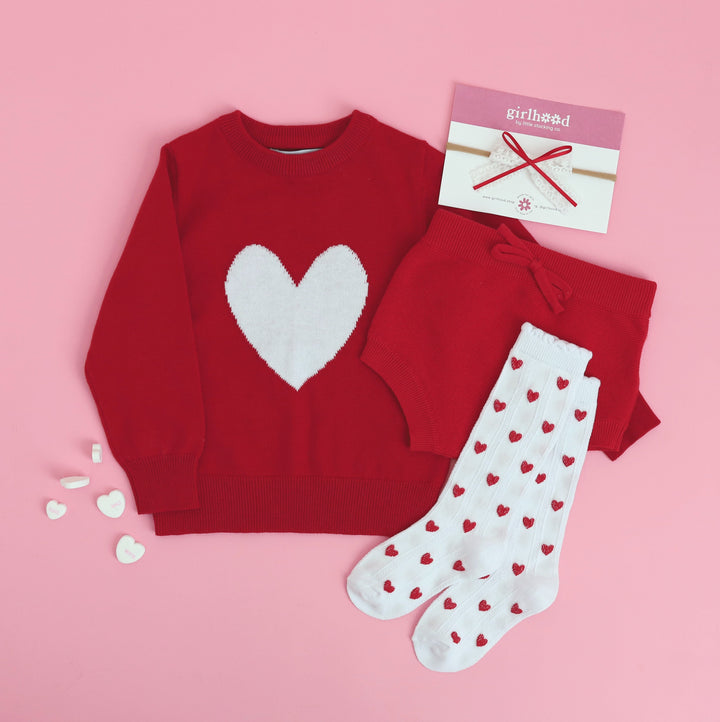 styled Valentine's flat lay featuring red baby sweater and bloomer set with matching heart knee highs and lace bow 