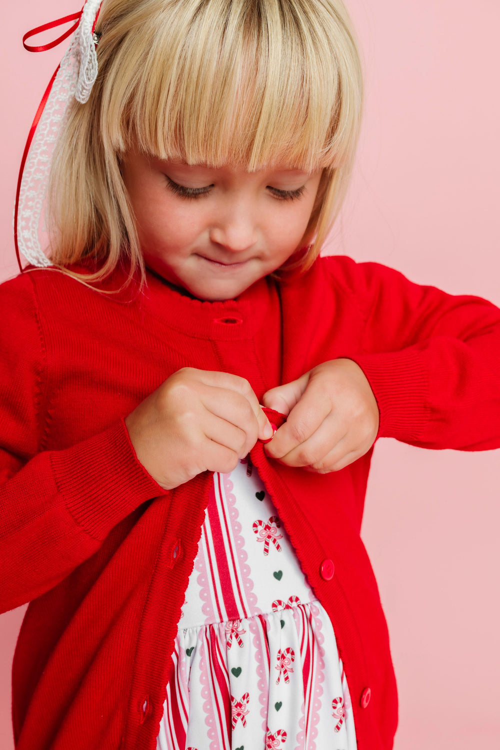 little girl buttoning bright red cotton knit sweater over candy cane print Christmas dress