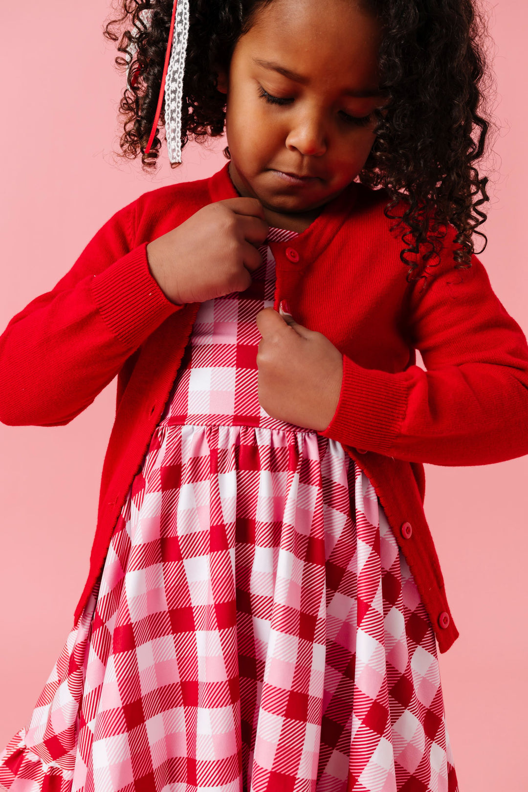 young girl buttoning bright red knit cardigan over plaid twirl dress in classic valentine's colors