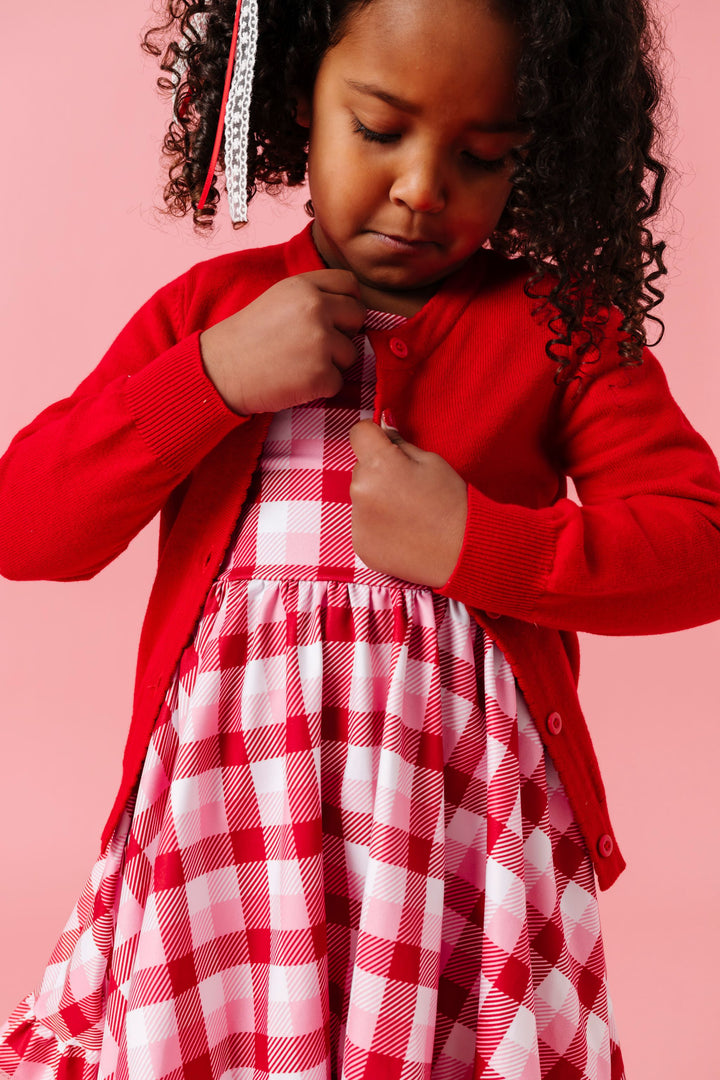 young girl buttoning bright red knit cardigan over plaid twirl dress in classic valentine's colors