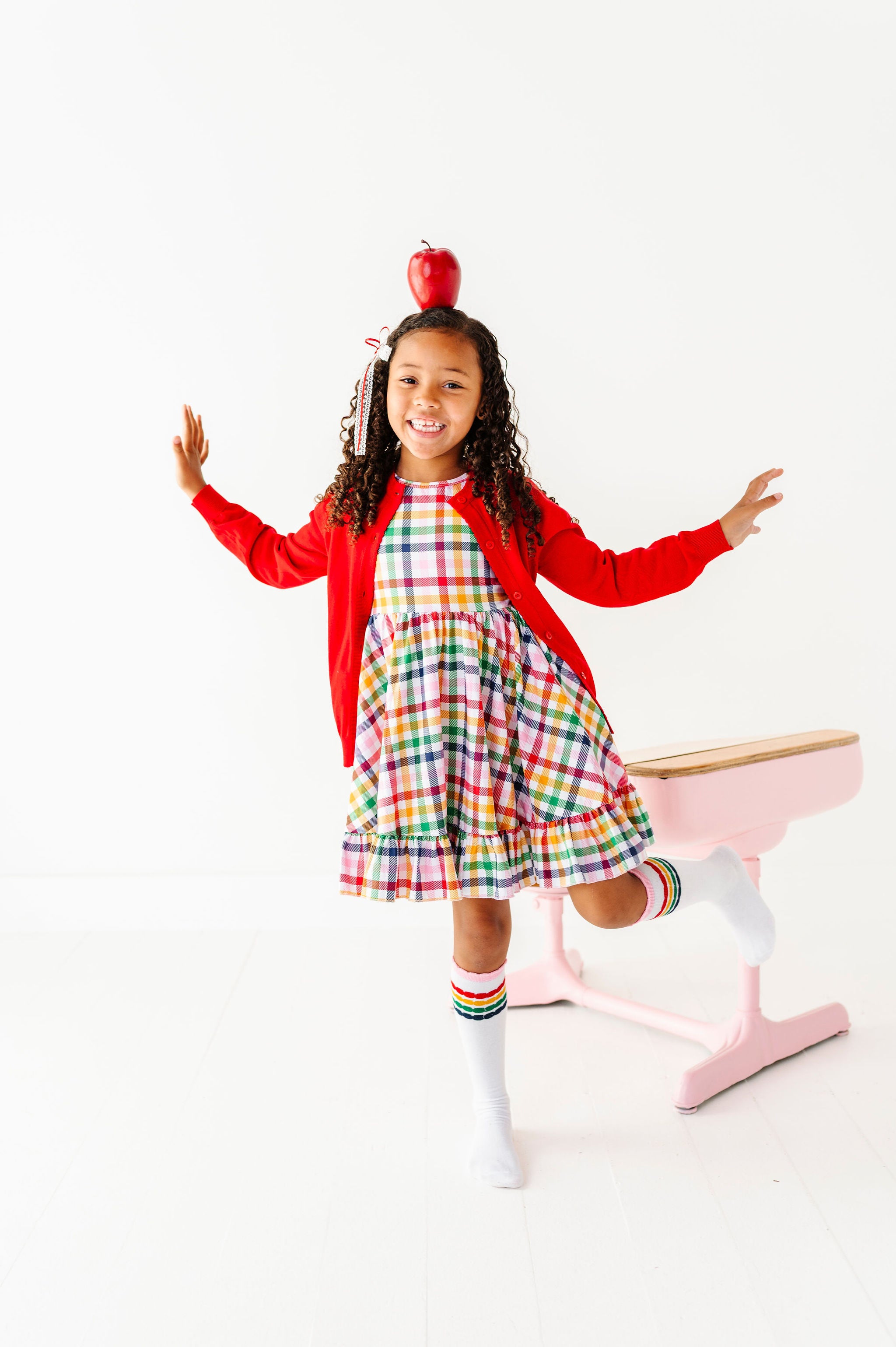 girl balancing apple on head in gingham print charm dress in primary colors paired with matching striped socks and bright red button front cardigan