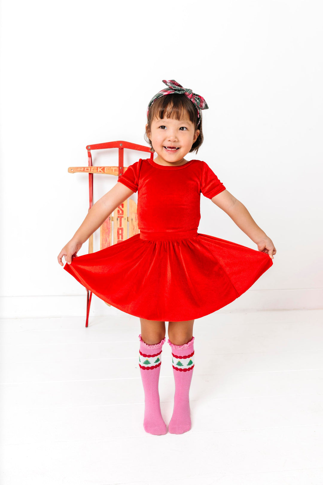 little girl in front of sled wearing matching bright red velvet top and skirt paired with pink tree knee highs and plaid headband for Christmas
