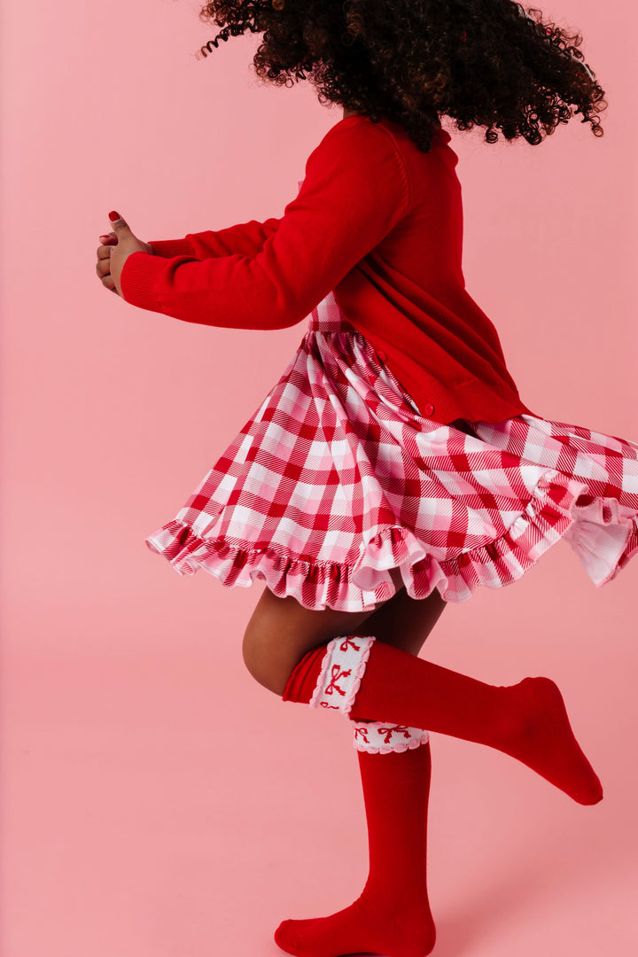 girl twirling in valentines inspired plaid twirl dress paired with red cotton knit cardigan and red knee highs with bow design