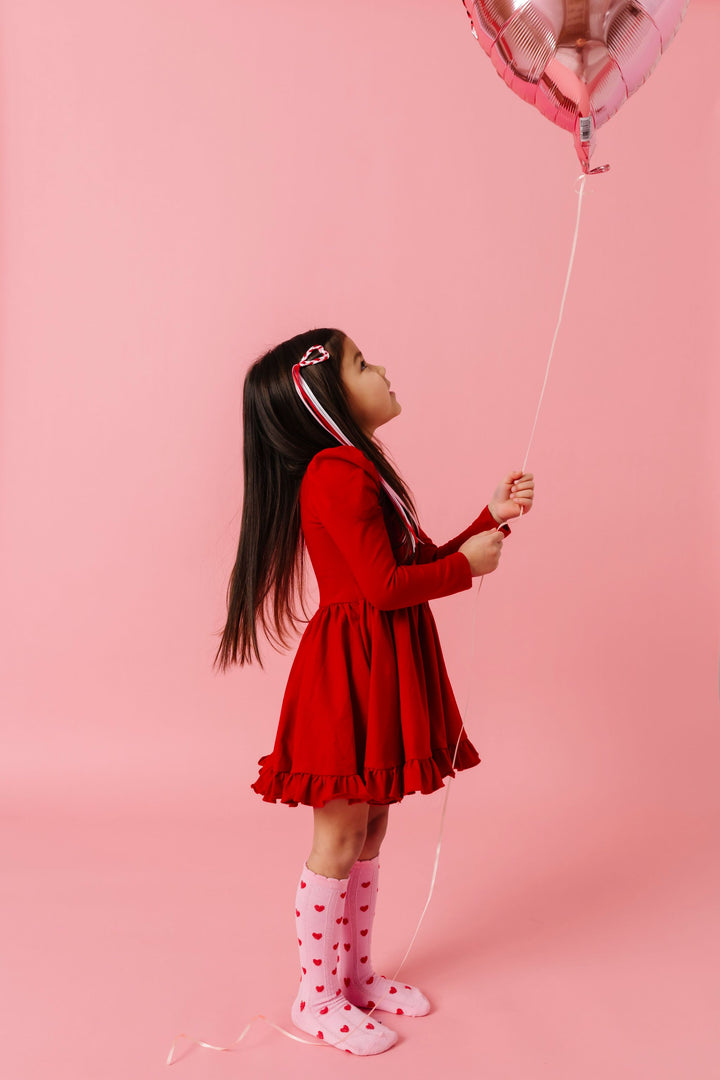 little girl holding heart balloon in bright red long sleeve twirl dress paired with pink scallop trimmed knee socks with little red hearts and a matching braided heart hair barrette with ribbon streamer  