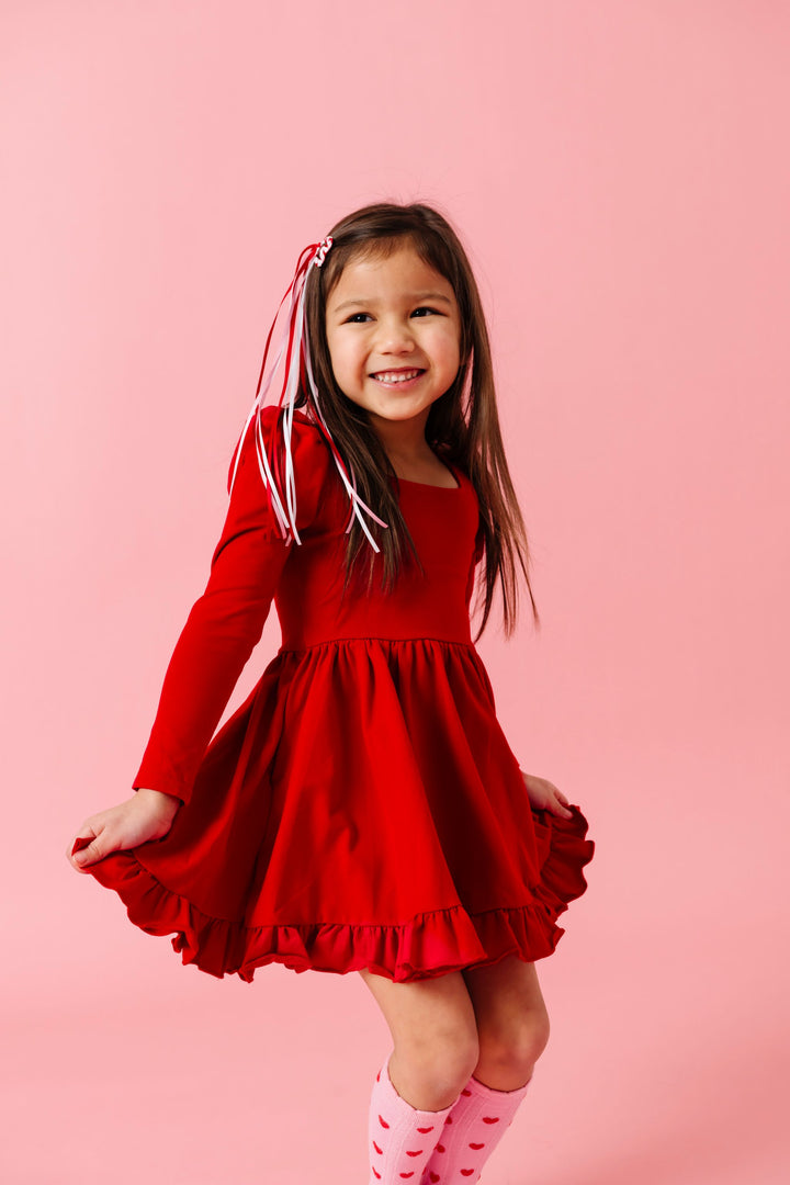 little girl in bright red long sleeve cotton blend twirl dress paired with valentines day heart hair clip with ribbon streamers and pink knee socks with red hearts