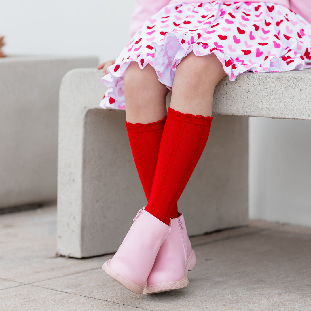 little girl sitting in bright red scallop trimmed knee high socks to match heart print dress for Valentine's Day