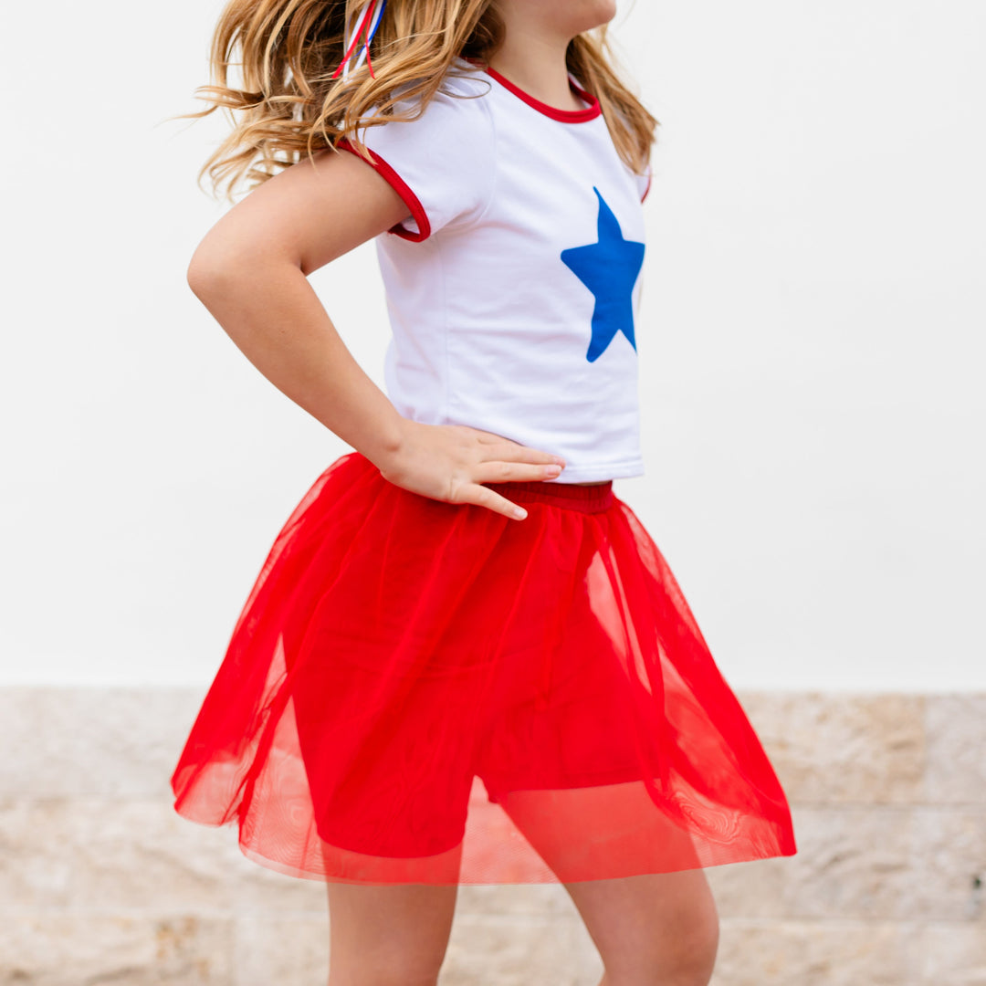 girl in white tee with red binding and blue star design paired with bright red tulle skirt with built in biker shorts for 4th of July