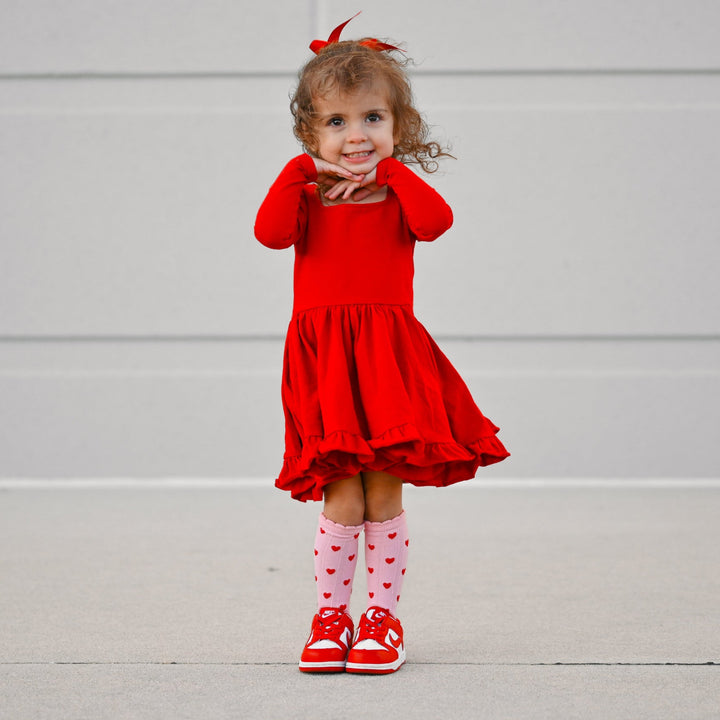 little girl posing in bright red long sleeve cotton blend twirl dress with matching red satin bow, pink scalloped knee socks with red heart design and red sneakers