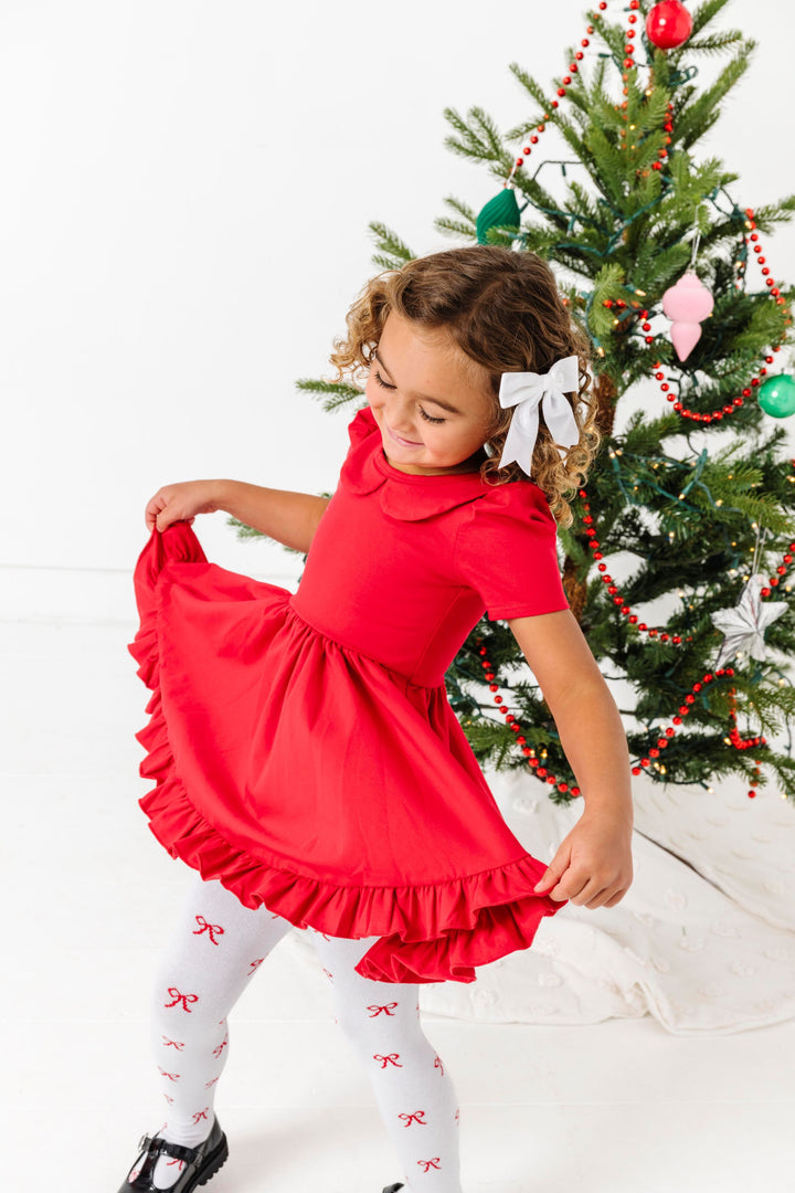 girl twirling by Christmas tree wearing bright red collared twirl dress pared with white velvet hair bow and white tights with red bow design