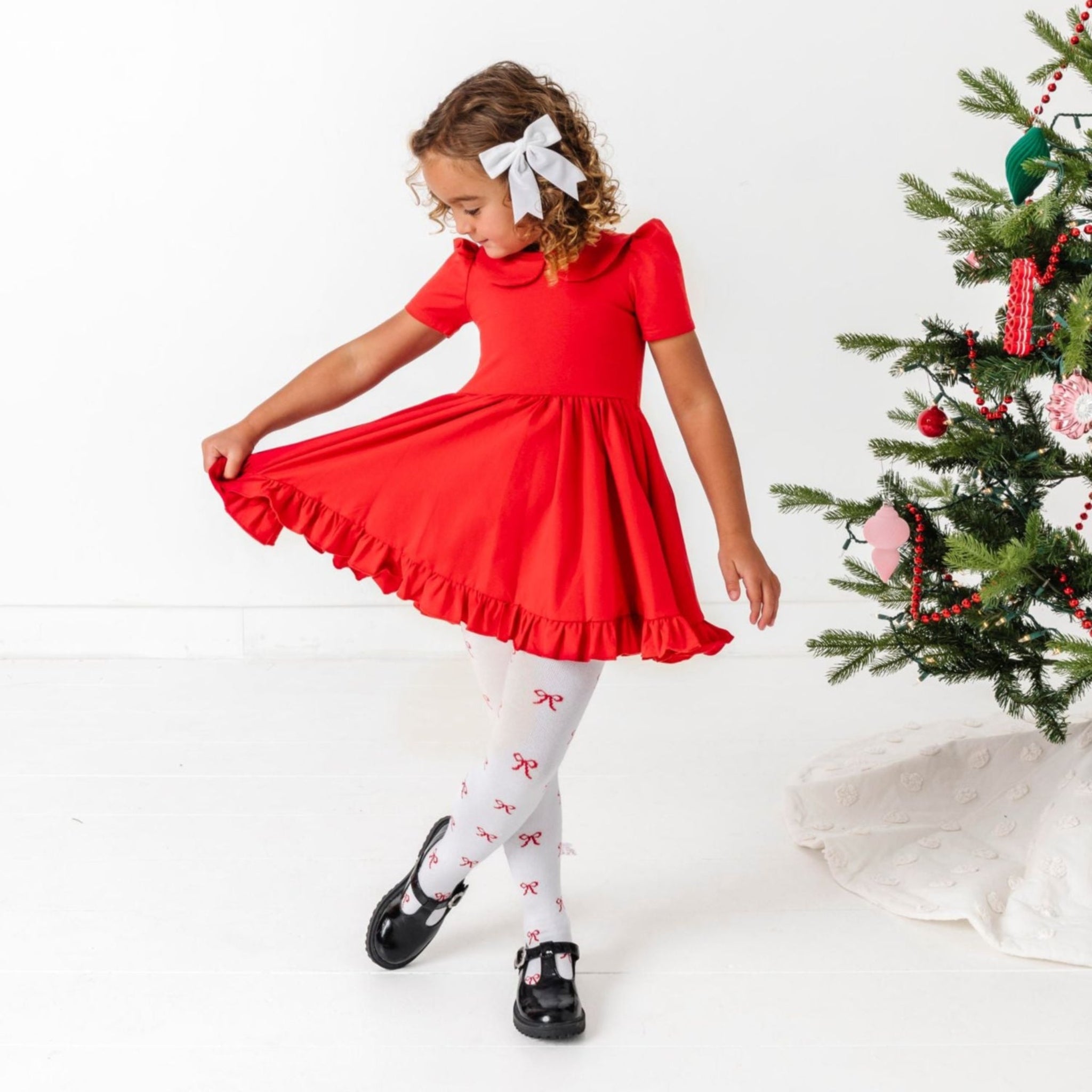 young girl in bright red short sleeve twirl dress with collar paired with white tights with red bow pattern and white velvet hair bow