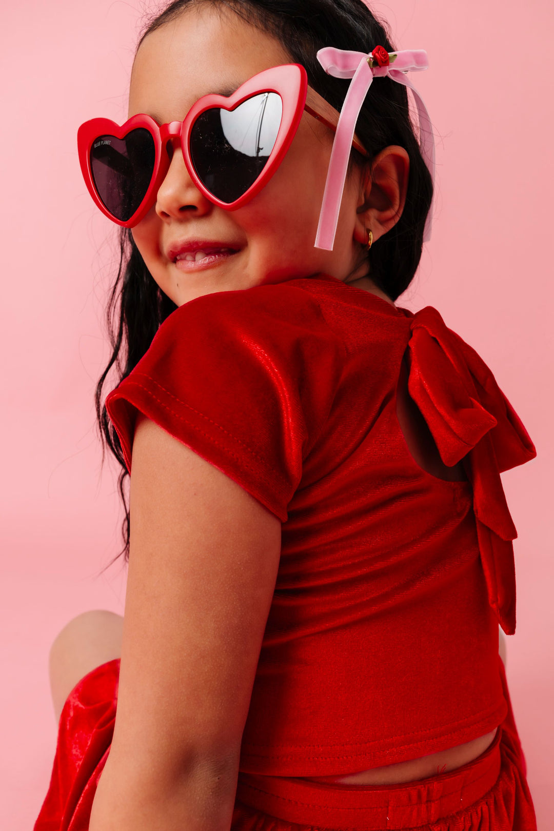 little girl looking over her shoulder in red heart sunglasses wearing bright red velvet top with bow ties at the back paired with pink velvet hair bow with red rosette 