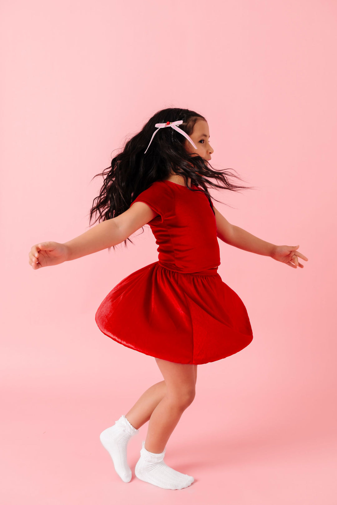 girl twirling in bright red velvet bow back top and skort set paired with white lace bobby socks and pink velvet rosette hair bow on clip