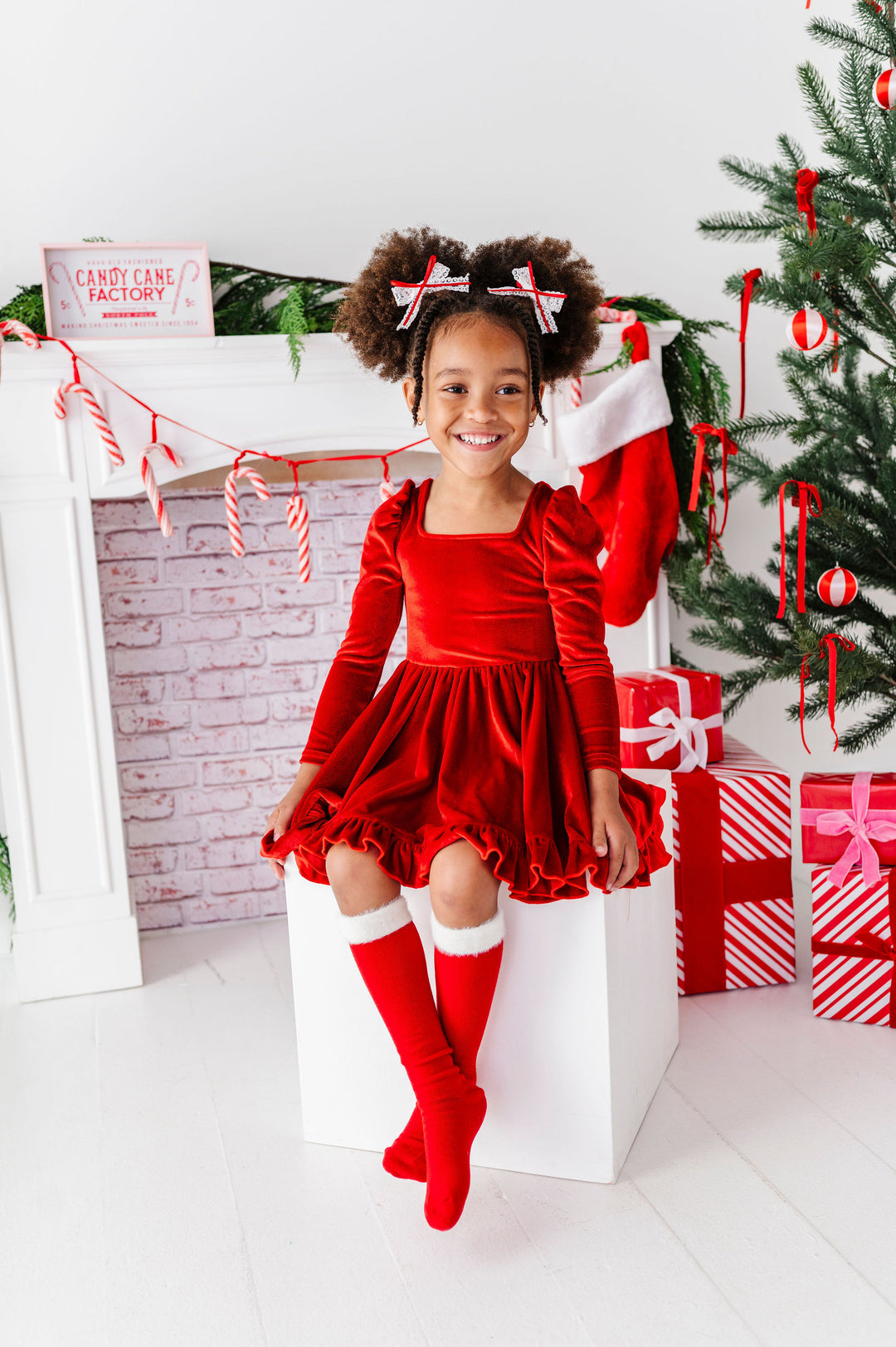 girl sitting in christmas scene wearing red velvet dress, santa boot knee high socks and matching hair bows