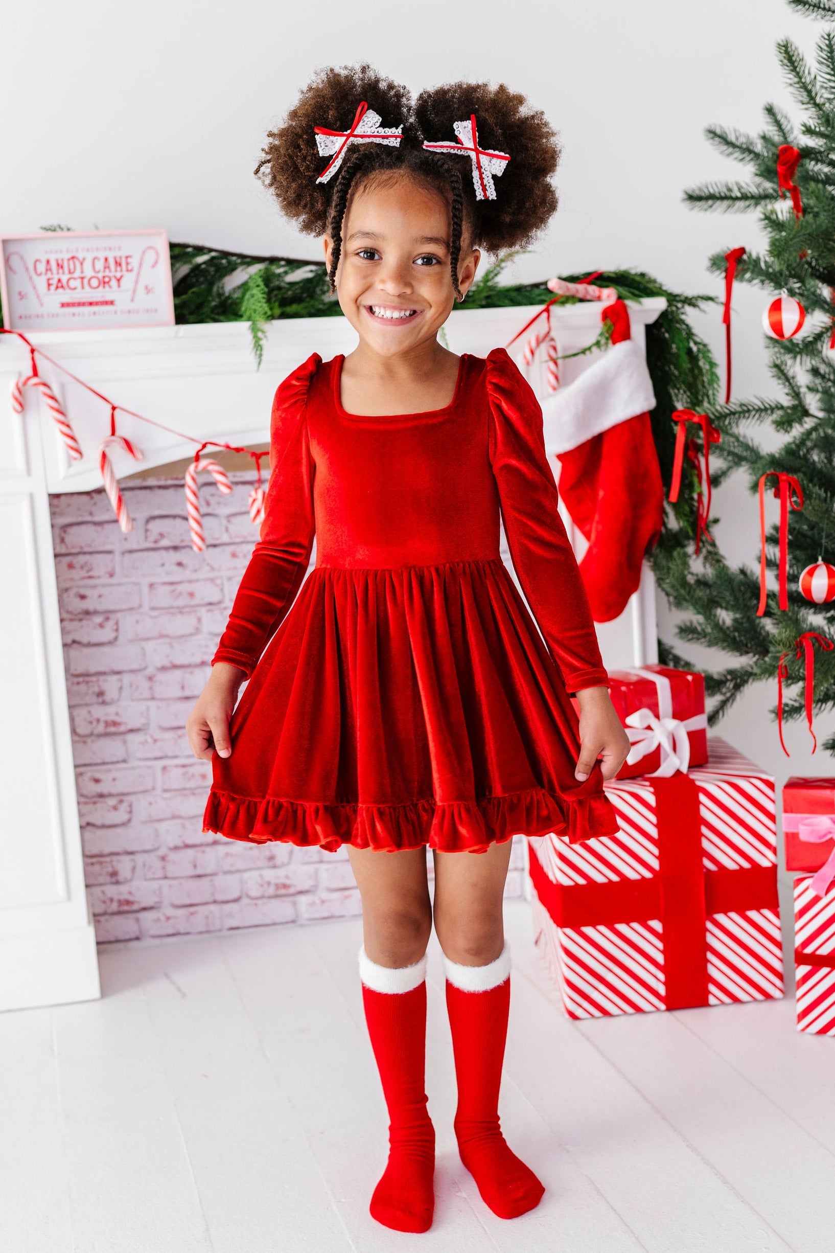 little girl wearing red velvet christmas dress with matching knee high socks and bows, standing in front of christmas tree with gifts
