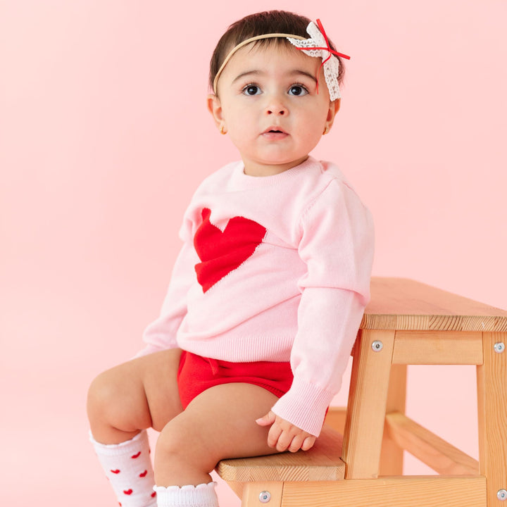 baby girl wearing pink Valentine's sweater with red heart, matching red sweater knit bloomers, lace bow on nylon headband and heart embroidered socks