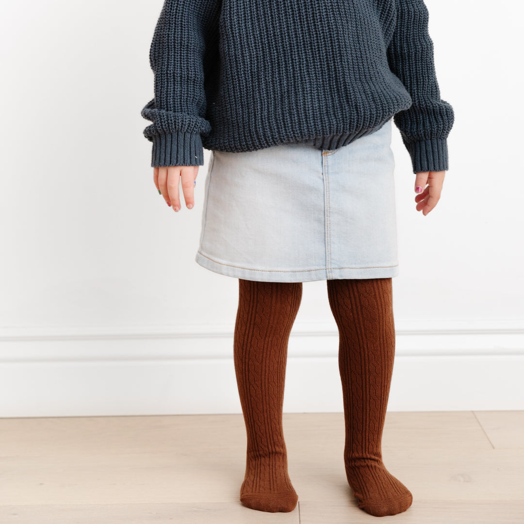 little girl standing in light denim skirt and brown cable knit tights