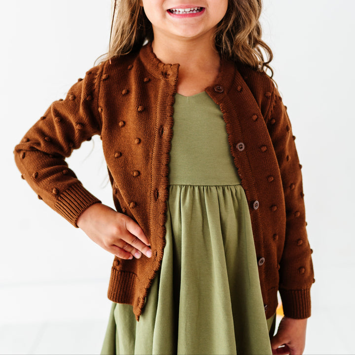 young girl in olive green v-neck twirl dress paired with brown cotton knit cardigan with raised bubble texture