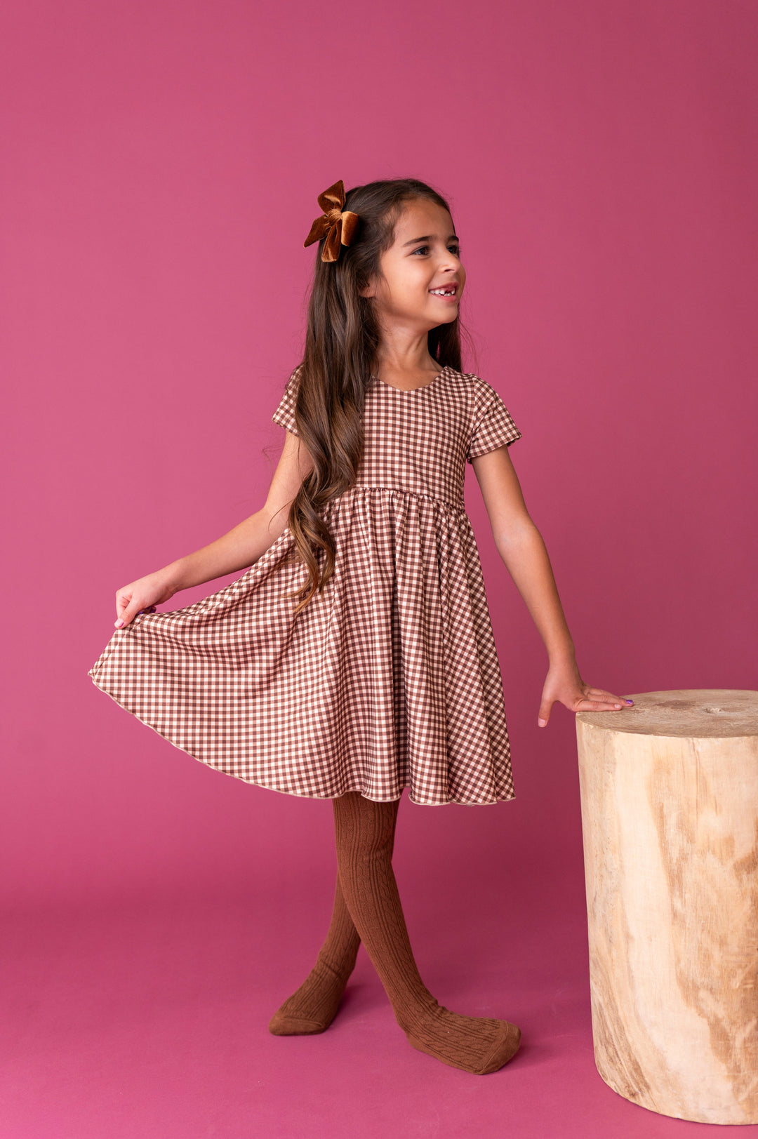 young girl in brown gingham twirl dress with v-shaped neckline paired with matching brown cable knit tights and velvet hair bow