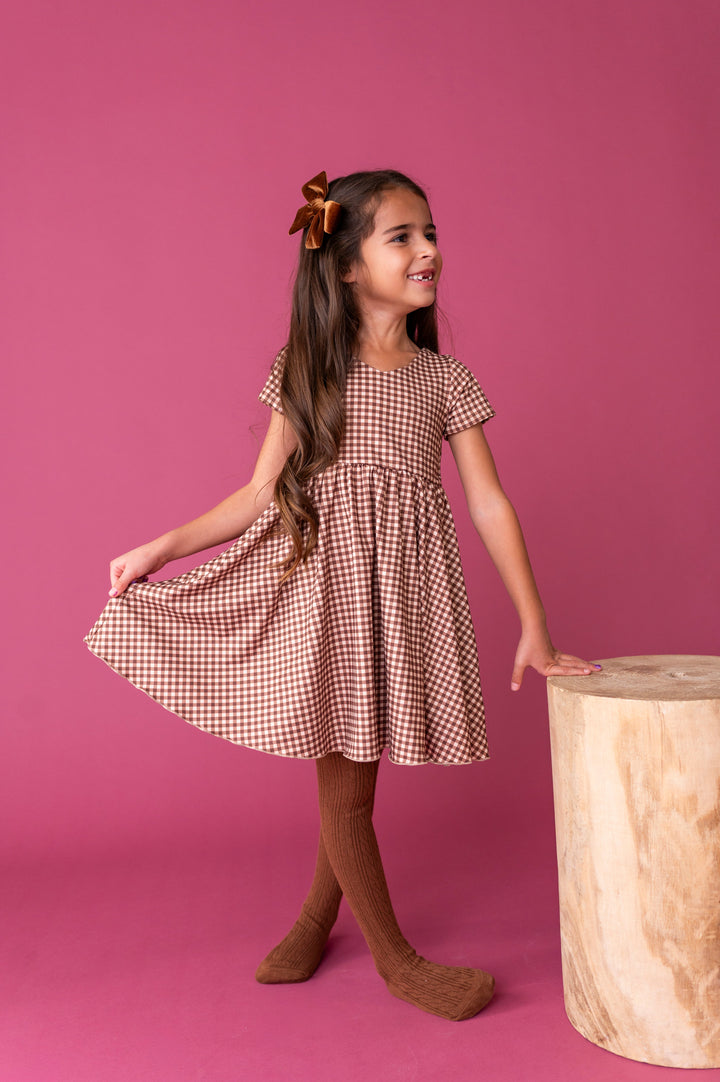 young girl in brown gingham twirl dress with v-shaped neckline paired with matching brown cable knit tights and velvet hair bow