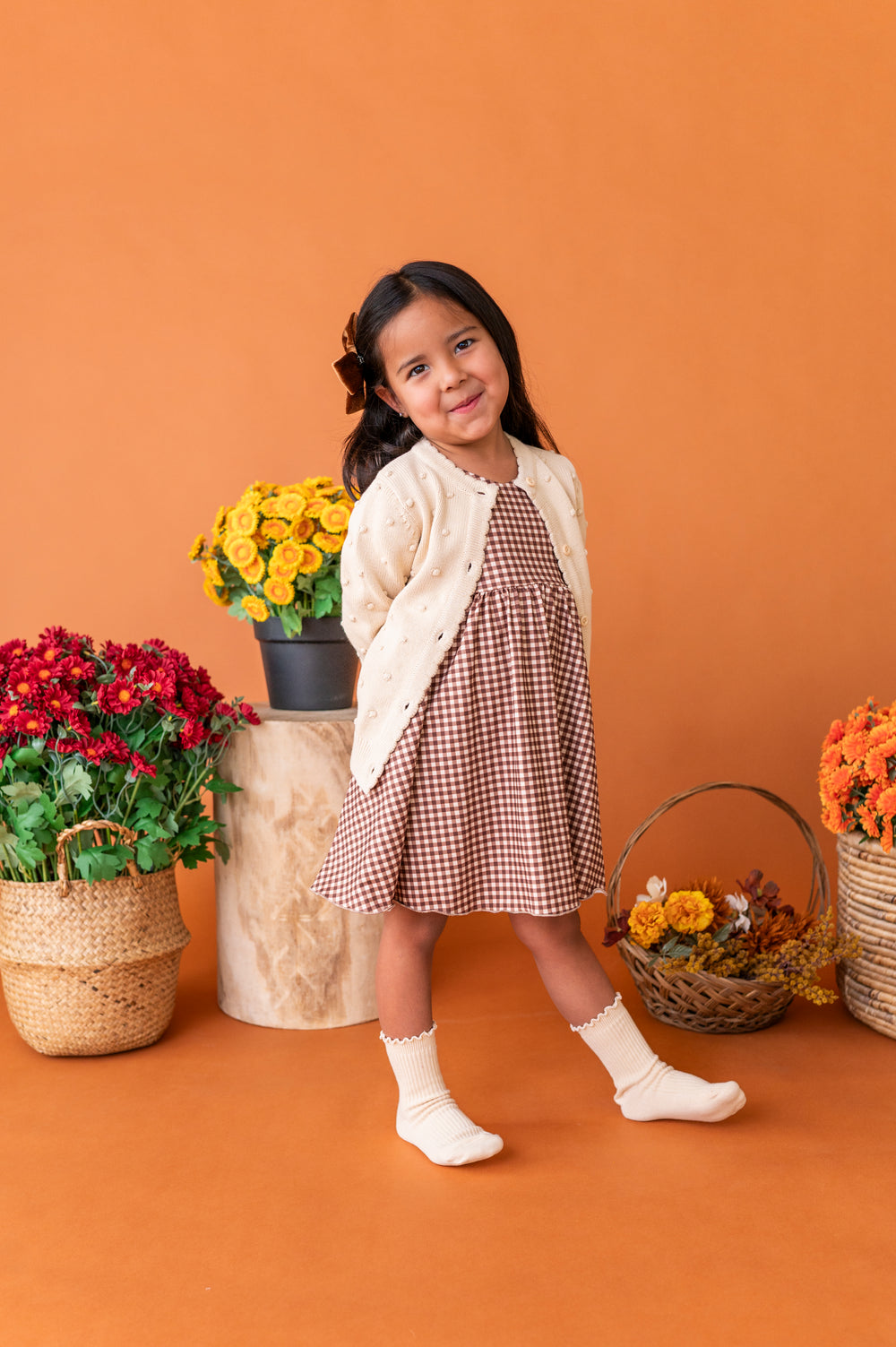 little girl in front of fall floral wearing brown gingham twirl dress paired with ream cardigan and ribbed midi socks