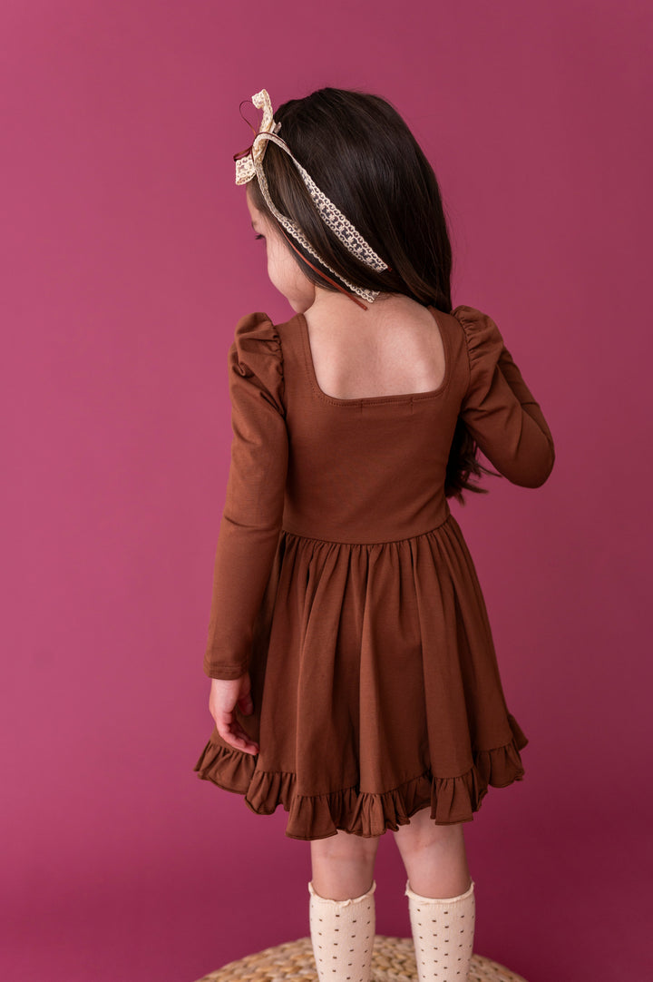 little girl showing back view of square neckline on brown long sleeve fall twirl dress paired with cream accessories