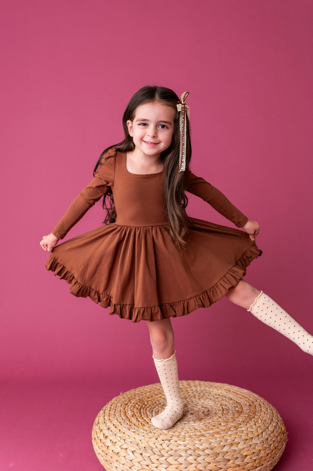 little girl balancing on woven cushion in rich brown long sleeve cotton blend twirl dress paired with cream dotted knee socks and lace bow