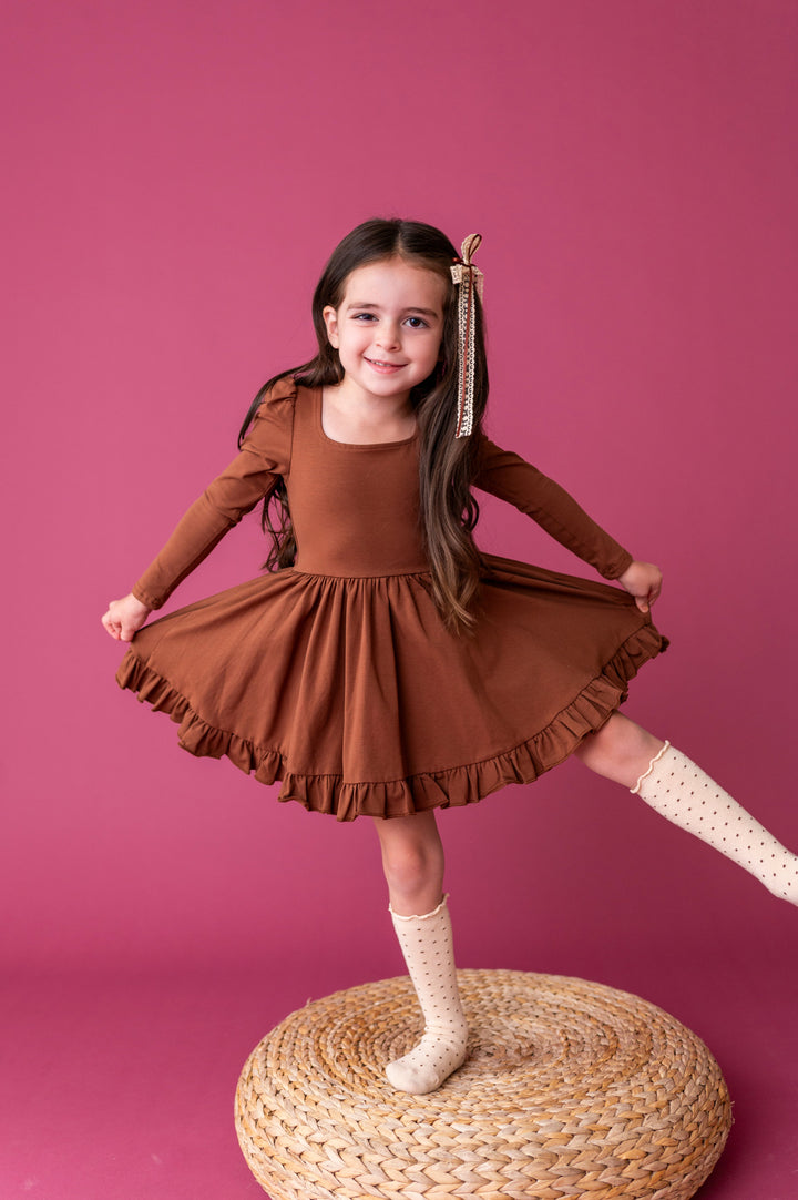 little girl balancing on woven cushion in rich brown long sleeve cotton blend twirl dress paired with cream dotted knee socks and lace bow