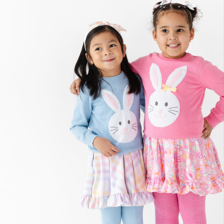 two girls in coordinating Easter outfits, one in a blue bunny sweater over a pastel gingham dress and one in a pink bunny sweater over a pink bunny and chick and egg dress