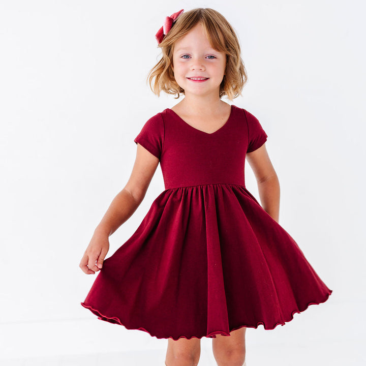 A young girl with blonde hair wearing a short-sleeved burgundy dress with a flared skirt, cream knee-high socks, and a matching red bow headband, standing against a white studio background.