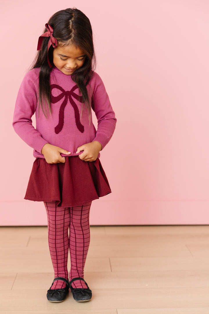 little girl in mulberry cotton knit pullover sweater with burgundy bow designh, burgundy cotton knit skort, matching hairbow and grid print tights