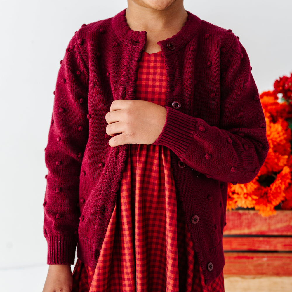closeup of little girl in burgundy cotton knit cardigan with raised bubble design paired with rust gingham twirl dress