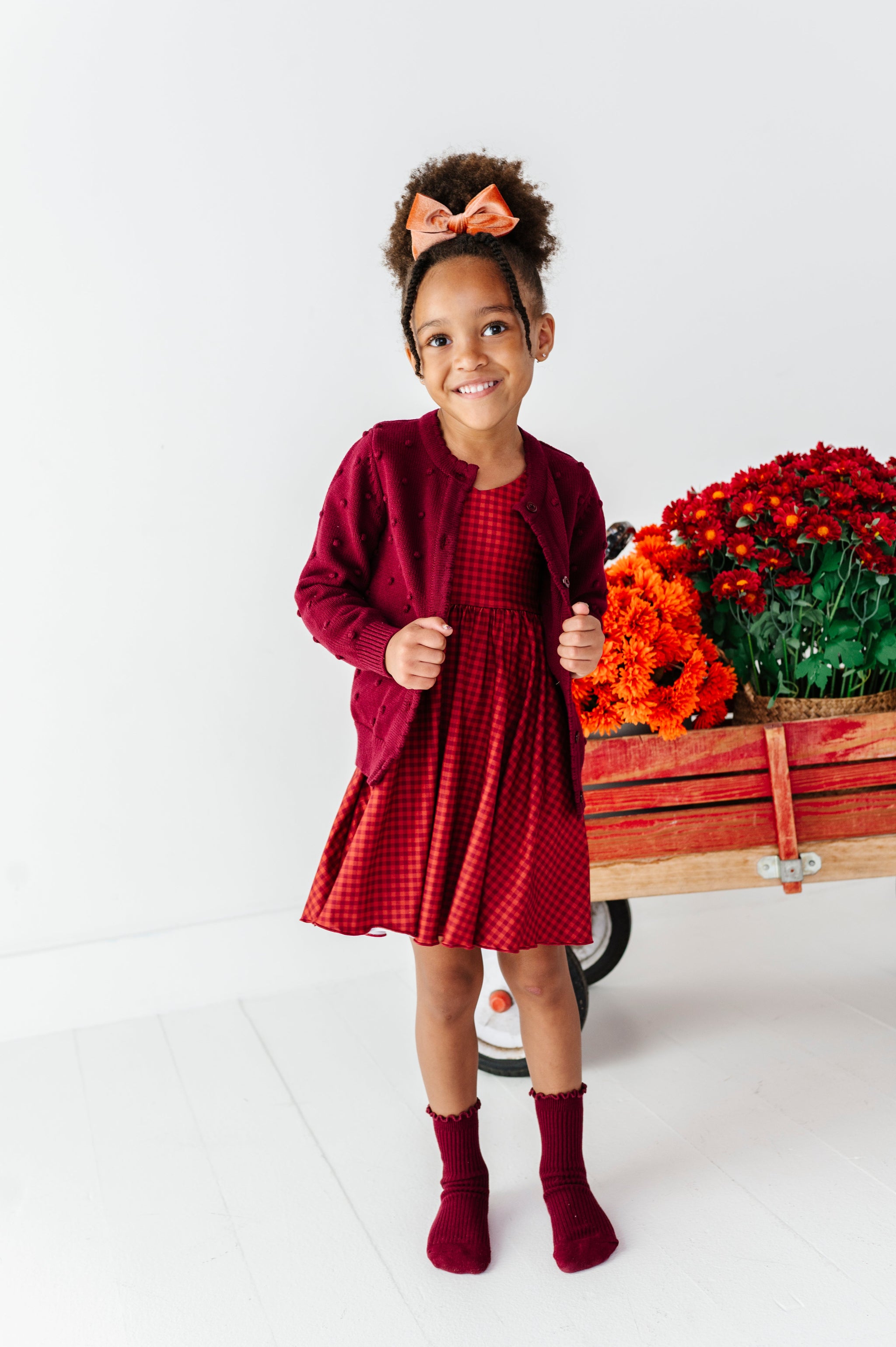 little girl posing in front of fall flower wagon in rust gingham twirl dress paired with burgundy bubble knit sweater and matching midi socks