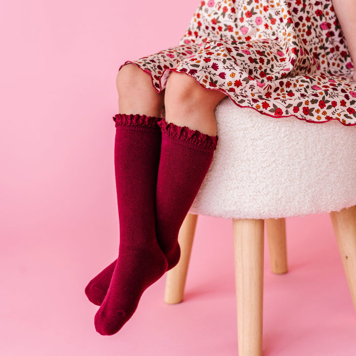 little girl sitting on fluffy white stool wearing burgundy lace top knee socks paired with fall floral dress with rust lettuce trim