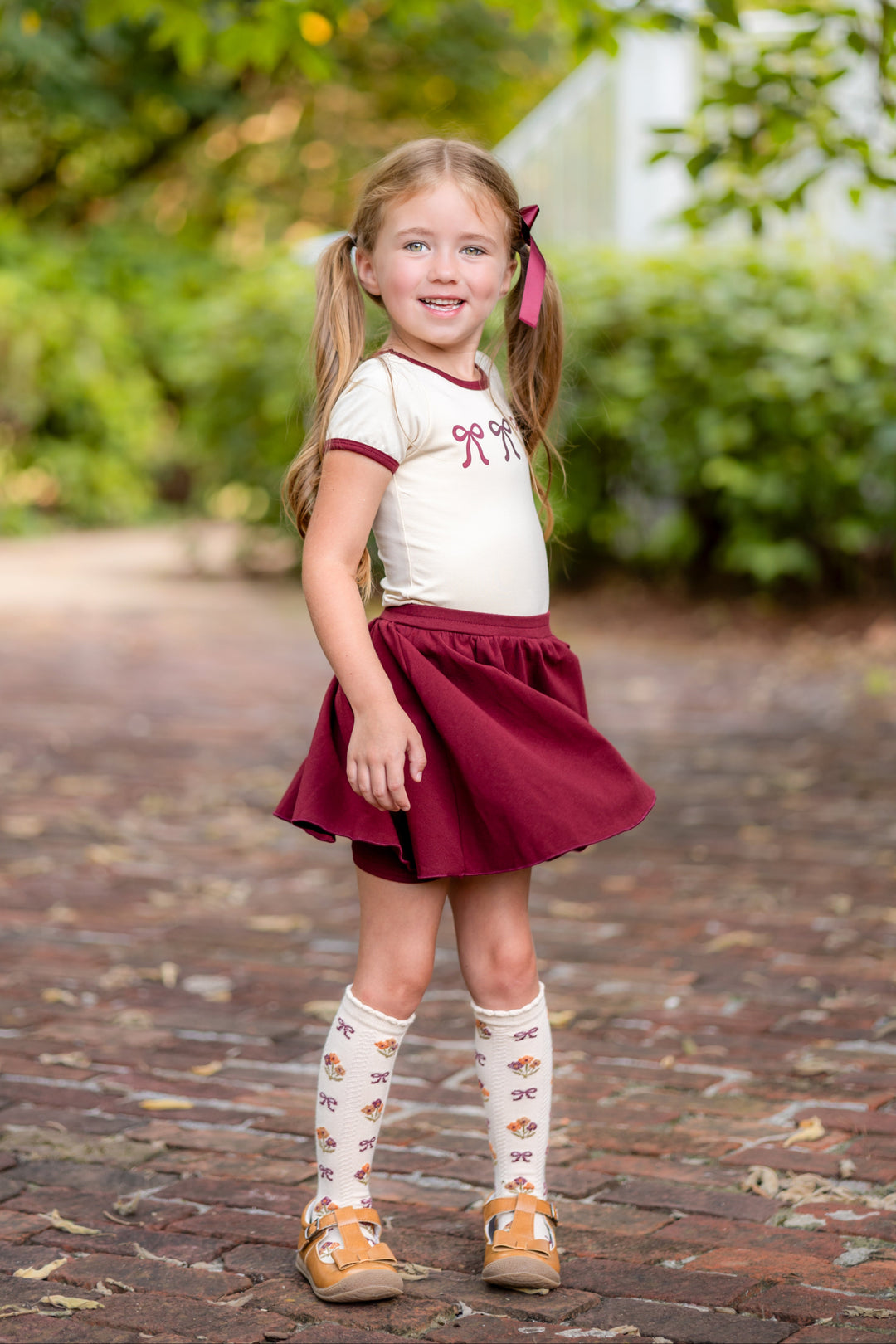 little girl in cream top with bow print and matching burgundy skort with built in shorts and matching knee highs and bow