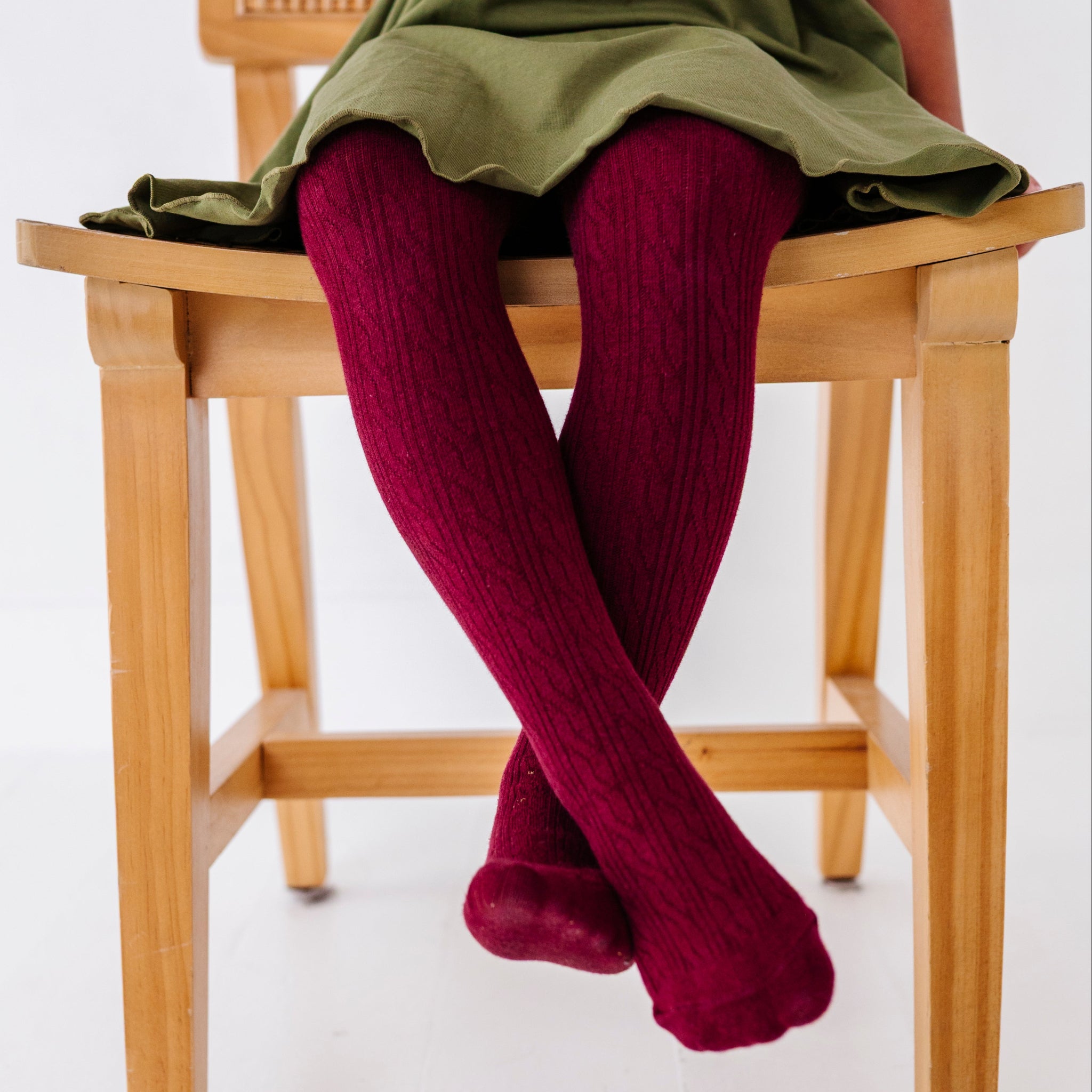 little girl sitting on wooden chair in burgundy cable knit tights paired with olive green lettuce trim twirl dress