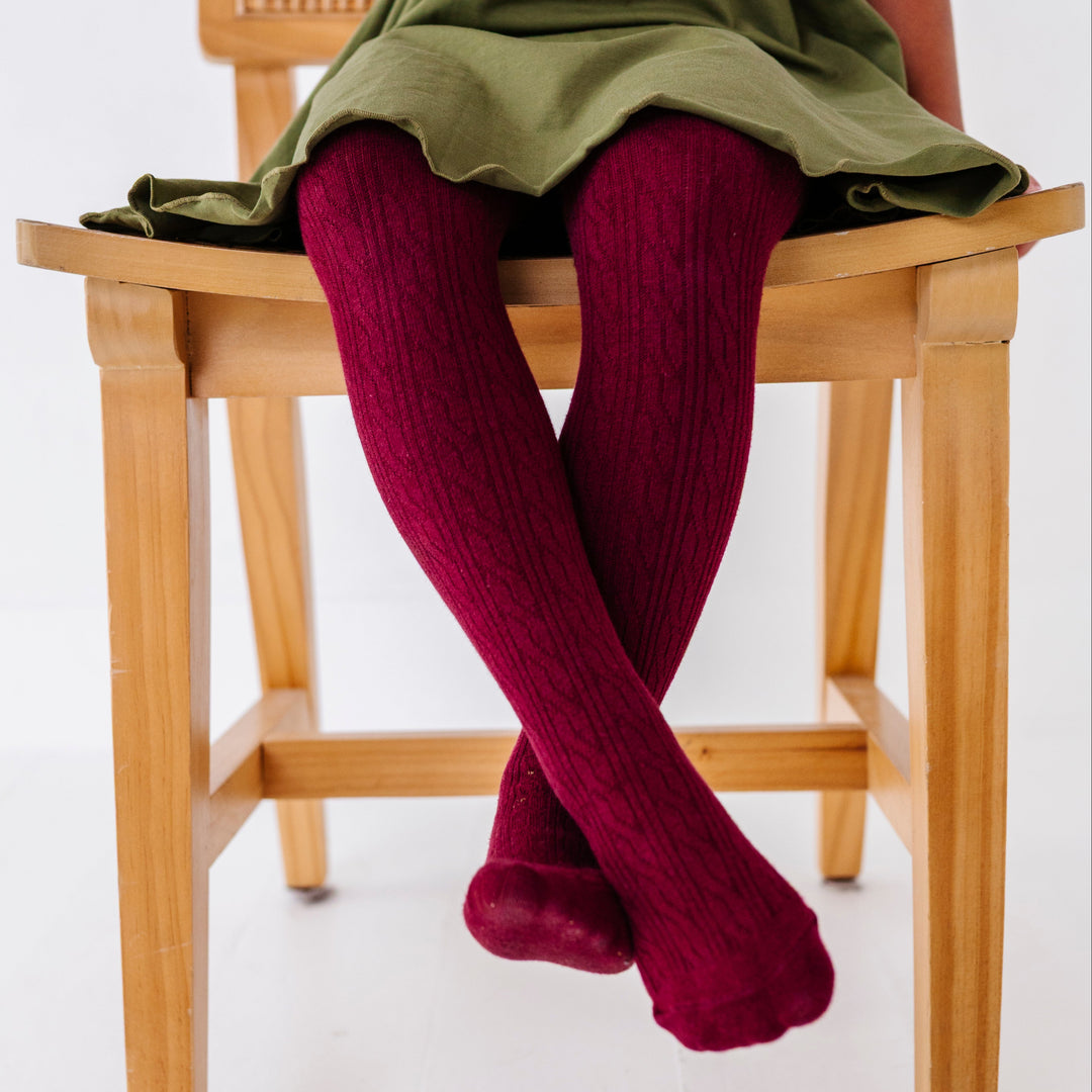 little girl sitting on wooden chair in burgundy cable knit tights paired with olive green lettuce trim twirl dress