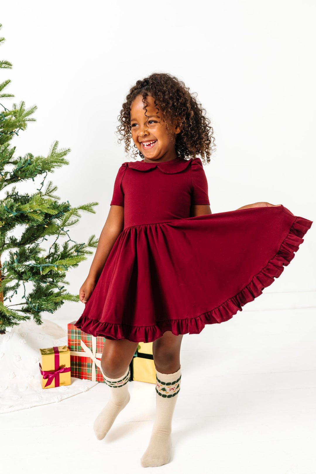 little girl by Christmas tree in burgundy collared twirl dress paired with cream fair isle print knee high socks 