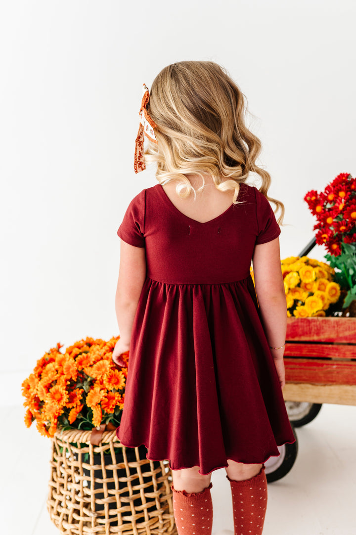little girl in front of fall flowers showing back view of v-neck design on burgundy cotton blend fall twirl dress