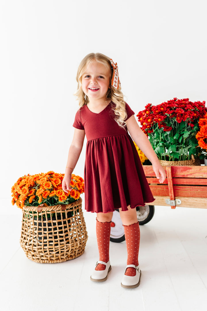 little girl in front of wagon full of fall flowers in burgundy cotton blend twirl dress with v-neck design and lettuce trim hem paired with rust dotted knee socks and floral hair bow