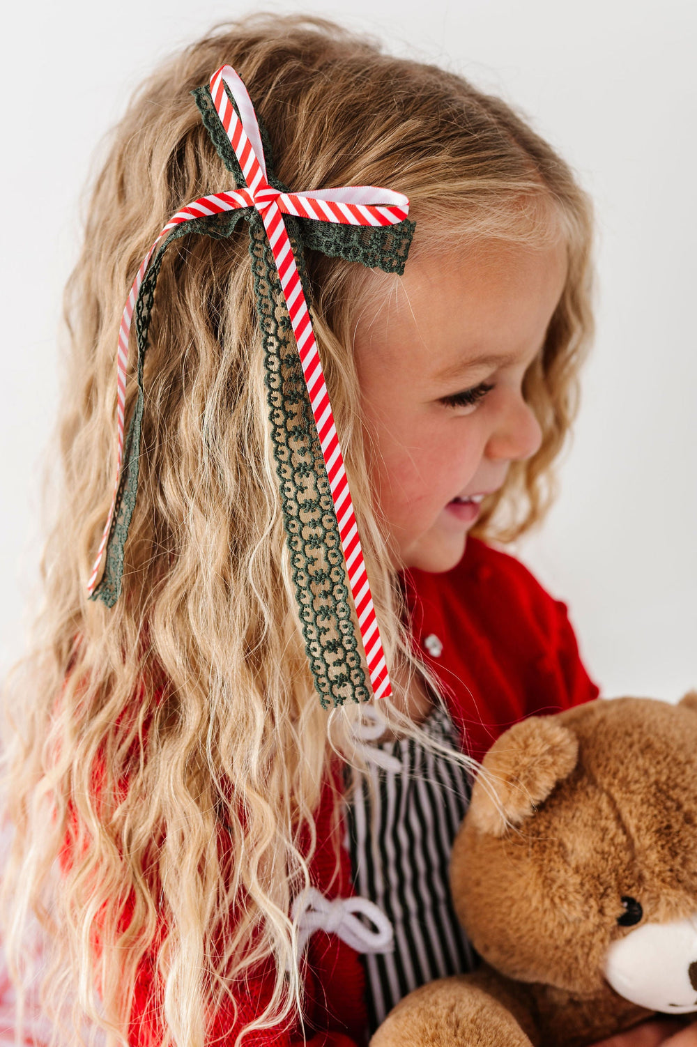 little girl holding teddy bear with candy cane striped and green lace hair bow in her hair