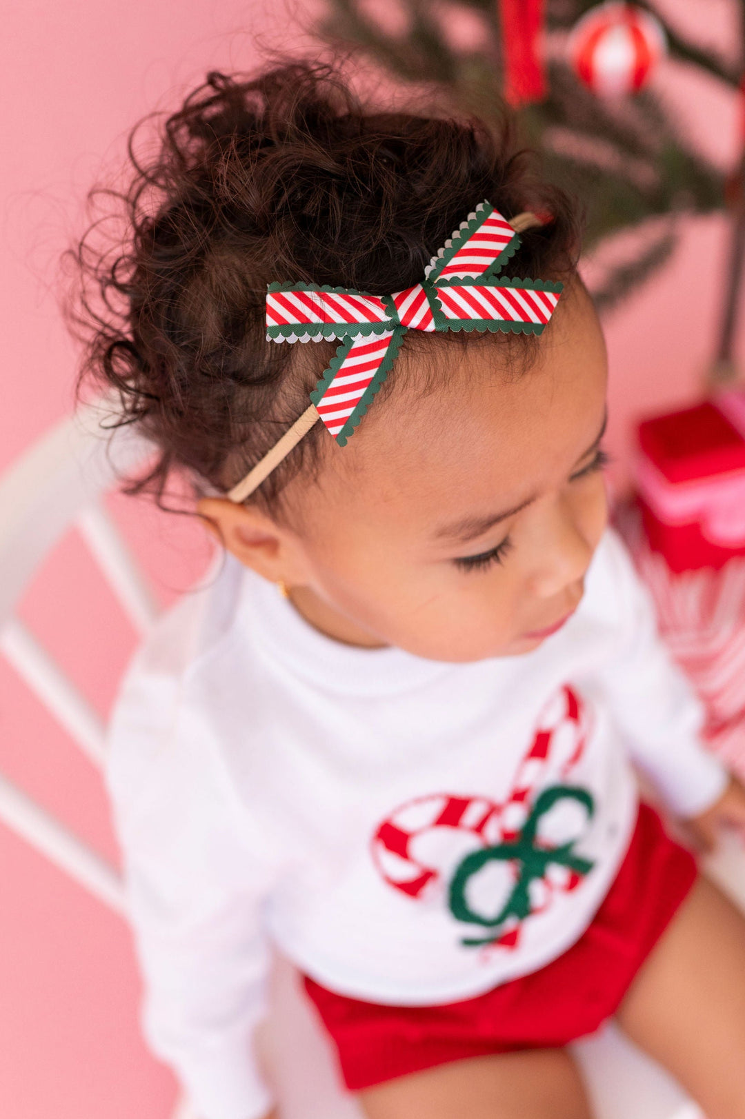toddler wearing a candy cane stripe baby bow with green scalloped edges on a nylon headband sitting on white chair and wearing a festive candy cane knitwear outfit