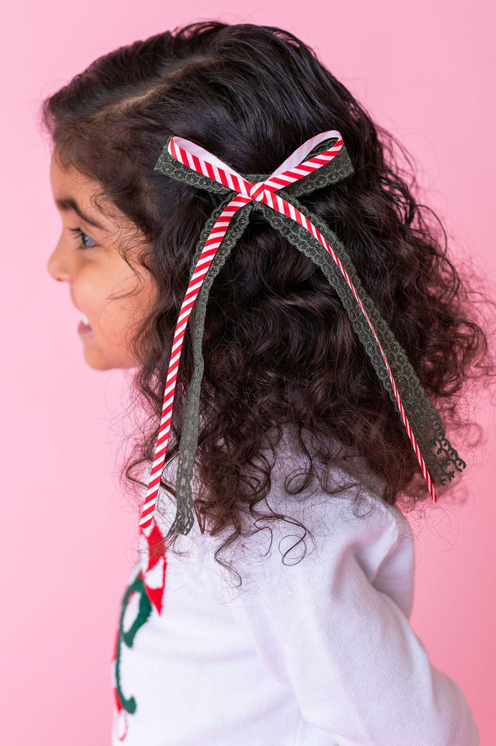 little girl with dark curly hair and white sweater wearing a candy cane stripe and green lace hair bow in her hair.