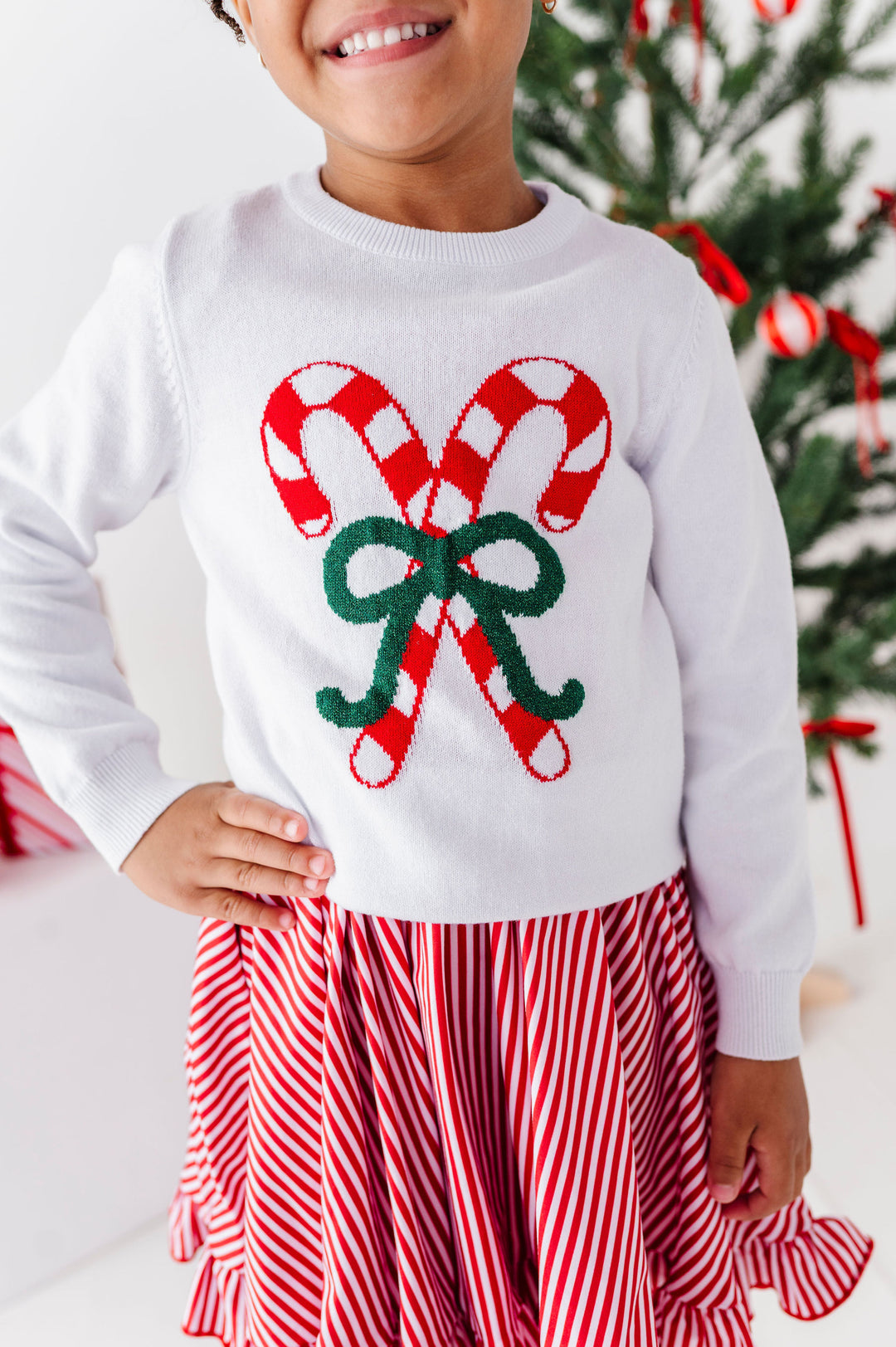 A child wearing a white long-sleeve sweater with a large red-and-white candy cane and green bow graphic on the front, paired with a red-and-white striped skirt, standing in front of a decorated Christmas tree.