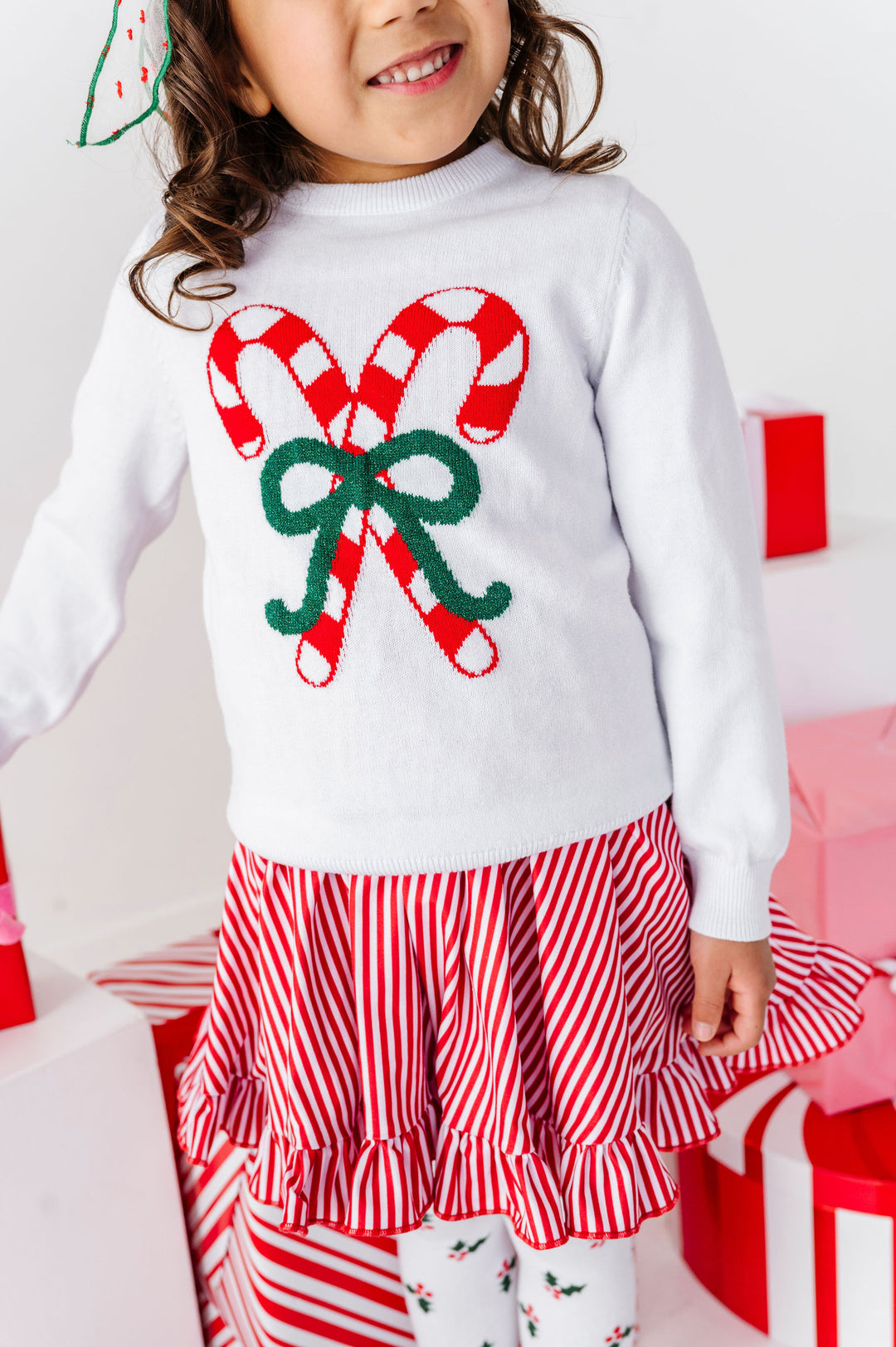A child wearing a white long-sleeve sweater with a large red-and-white candy cane and green bow graphic on the front, paired with a red-and-white striped ruffled skirt and matching striped tights, standing among pink and red gift boxes.