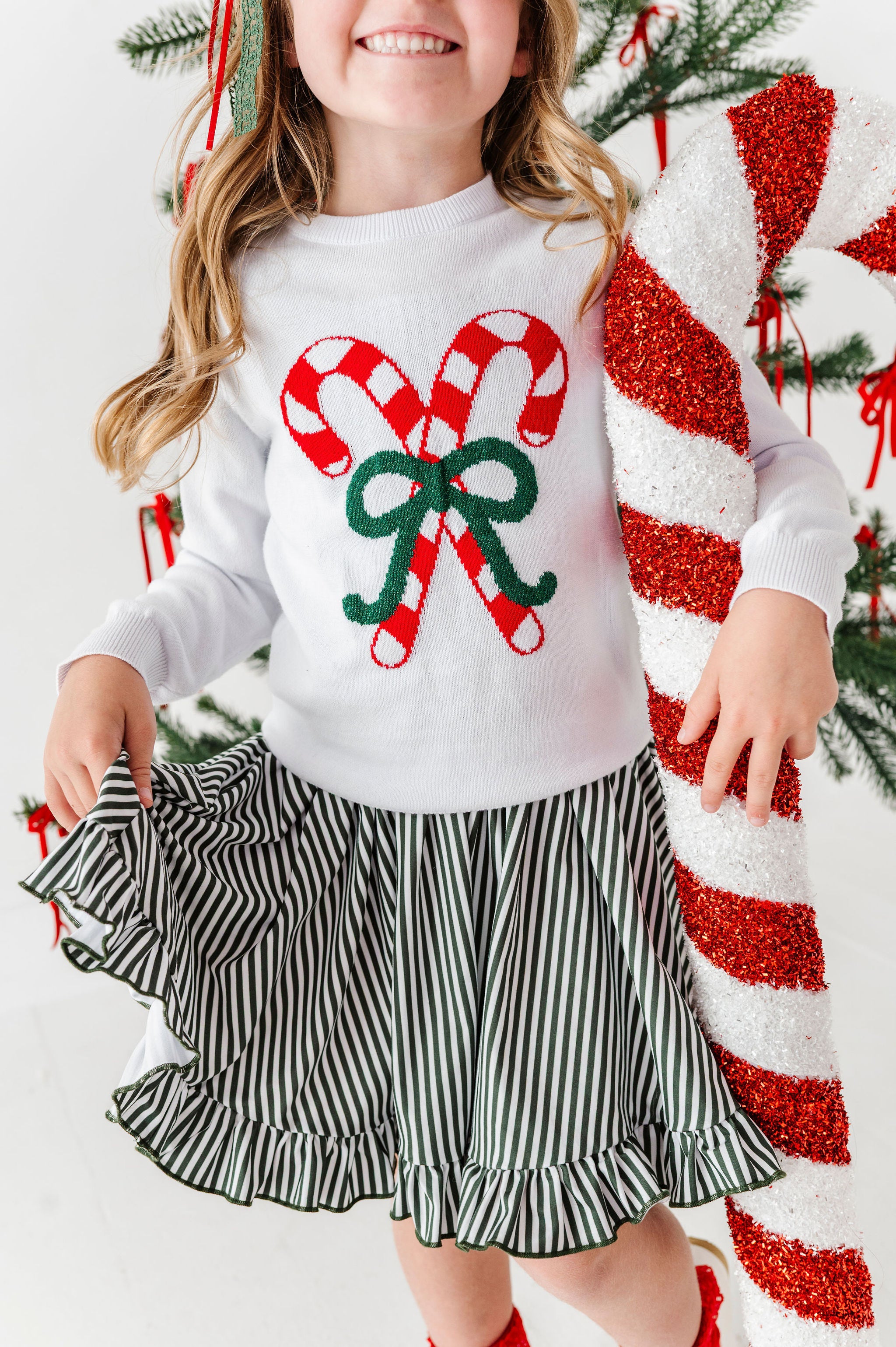little girl wearing white candy cane sweater over dark green pinstriped ruffle hemmed dress with one arm wrapped around a large candy cane decoration
