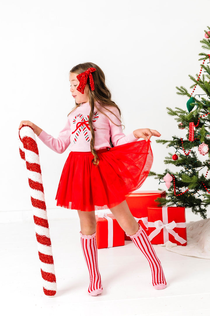 little girl holding candy cane decoration in pink candy cane sweater, red tutu skirt, sparkly red hair bow and matching pink and red striped socks