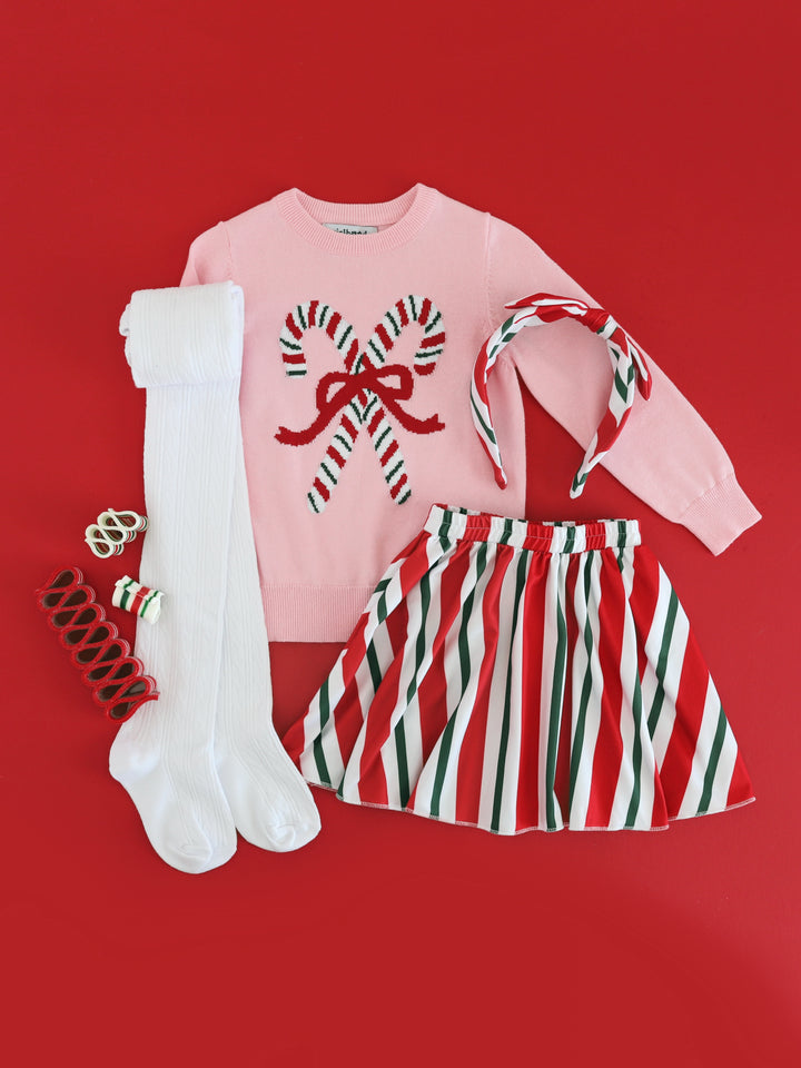 flat lay of girls' pink cotton knit sweater with red, white and green striped candy cane design paired with matching striped skirt and headband and white cable knit tights