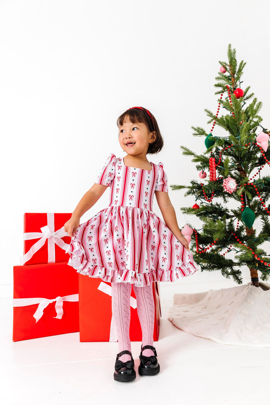 little girl in front of Christmas tree and gifts in vintage candy cane print twirl dress paired with pink open knit tights and black mary janes