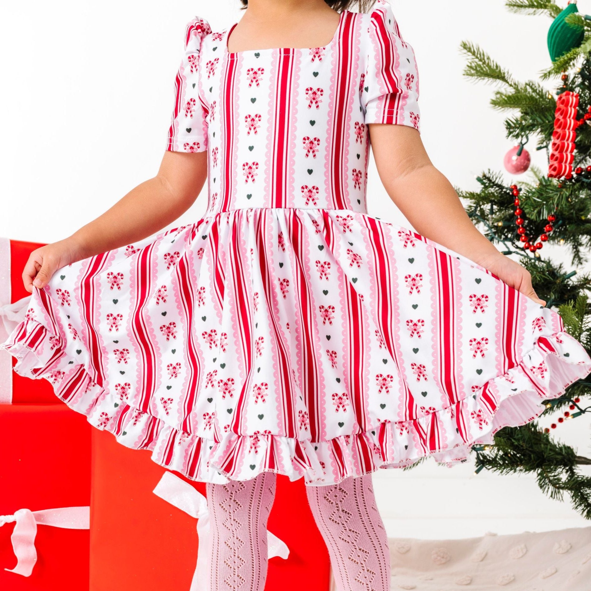 little girl by Christmas tree wearing candy cane themed twirl dress on white base with matching pink open knit tights